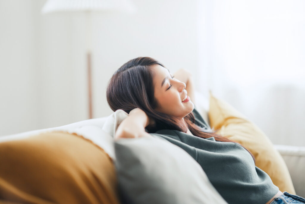 A woman sits back on her couch with her hands resting behind her head and her eyes closed in relaxation